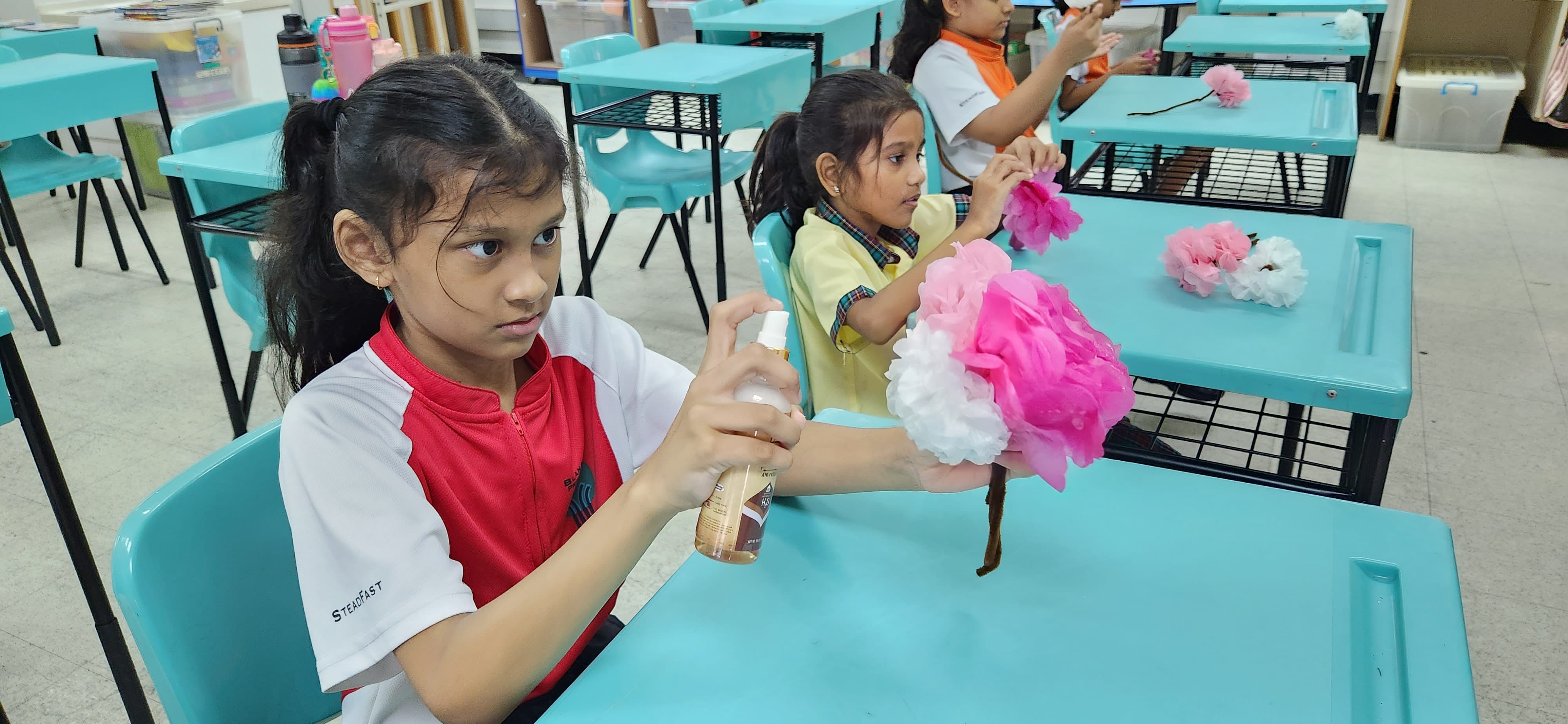 Girls in school uniforms sit at teal desks making pink and white tissue paper flowers with a spray bottle.